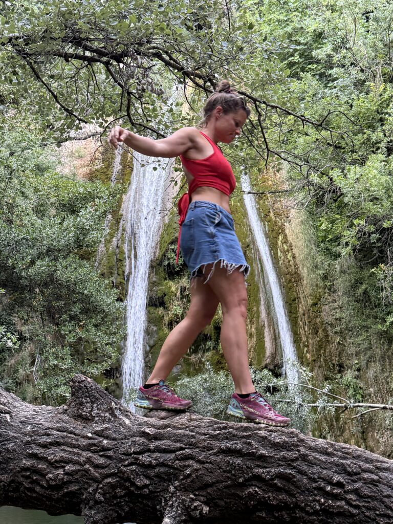 Marlen balanciert in rotem Top, Jeansrock und Turnschuhen über einen dicken Baumstamm, hinter ihr stürzt ein Wasserfall über eine bemooste Felswand.