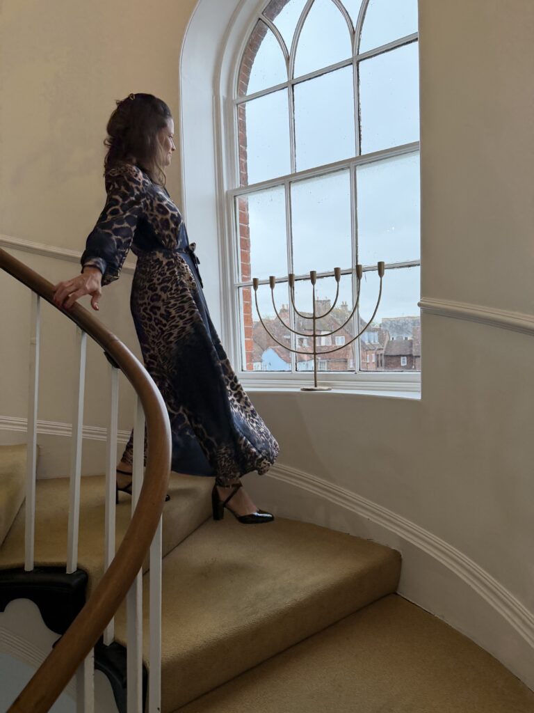 Marlen schreitet in einem langen Kleid mit Leopardenmuster eine geschwungene Treppe hinunter, die Hand am Holzgeländer, im hohen Fenster steht ein Kerzenleuchter.