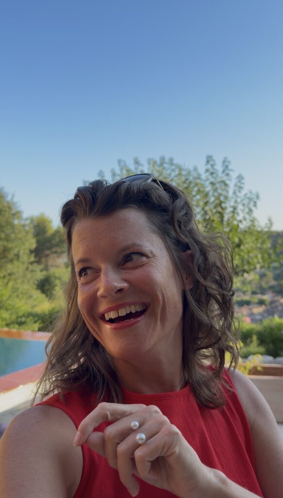 Marlen lacht herzhaft in einem roten Top, Perlenring an der Hand, hinter ihr Bäume, ein Pool und blauer Sommerhimmel.